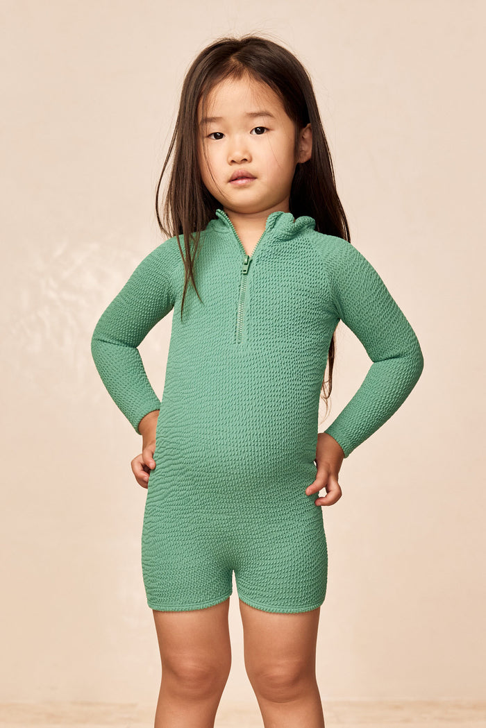 Front view of Hilo kids one-piece in Pond Crinkle with crinkle texture, long sleeves, front zipper and short legs on beige background
