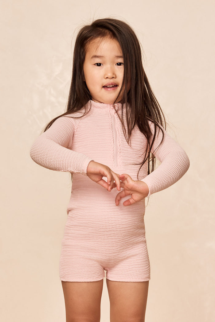 Front view of Hilo One Piece Kids in powder pink crinkle with long sleeves, center front zipper and scoop neckline on beige