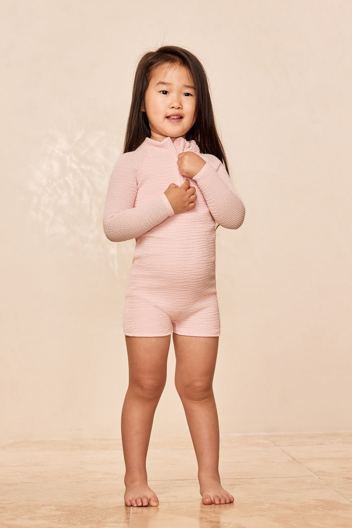 Three-quarter front of Hilo One Piece Kids in Powder Pink Crinkle, showing textured crinkle fabric, long sleeves and scoop neckline against a pale beige background