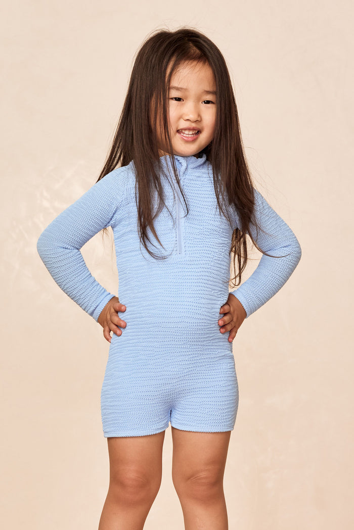 Front view of Hilo One Piece Kids in Sky Crinkle with long sleeves, crinkle texture and short leg on beige backdrop