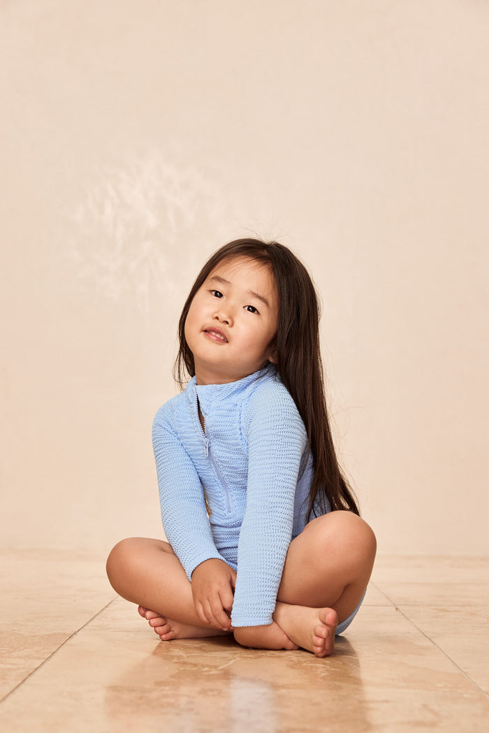 Child sitting on floor wearing Hilo One Piece Kids in Sky Crinkle, showing long sleeves and center-front zipper on beige backdrop