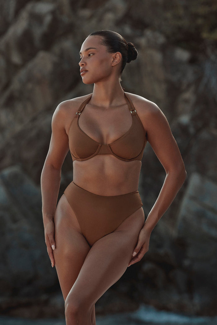 Three-quarter view of Honolulu Top in caramel on a model, showcasing underwire cups and halter straps with a rocky beach background