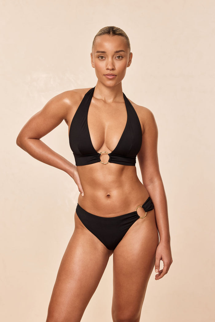 Front view of Ibiza Top in black showcasing plunging halter neckline, wide underband and gold ring detail on beige background