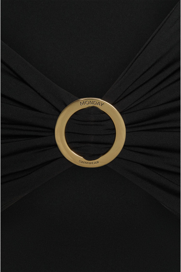 Gold circular buckle engraved 'MONDAY' and 'SWIMWEAR' on a black ruched swimsuit