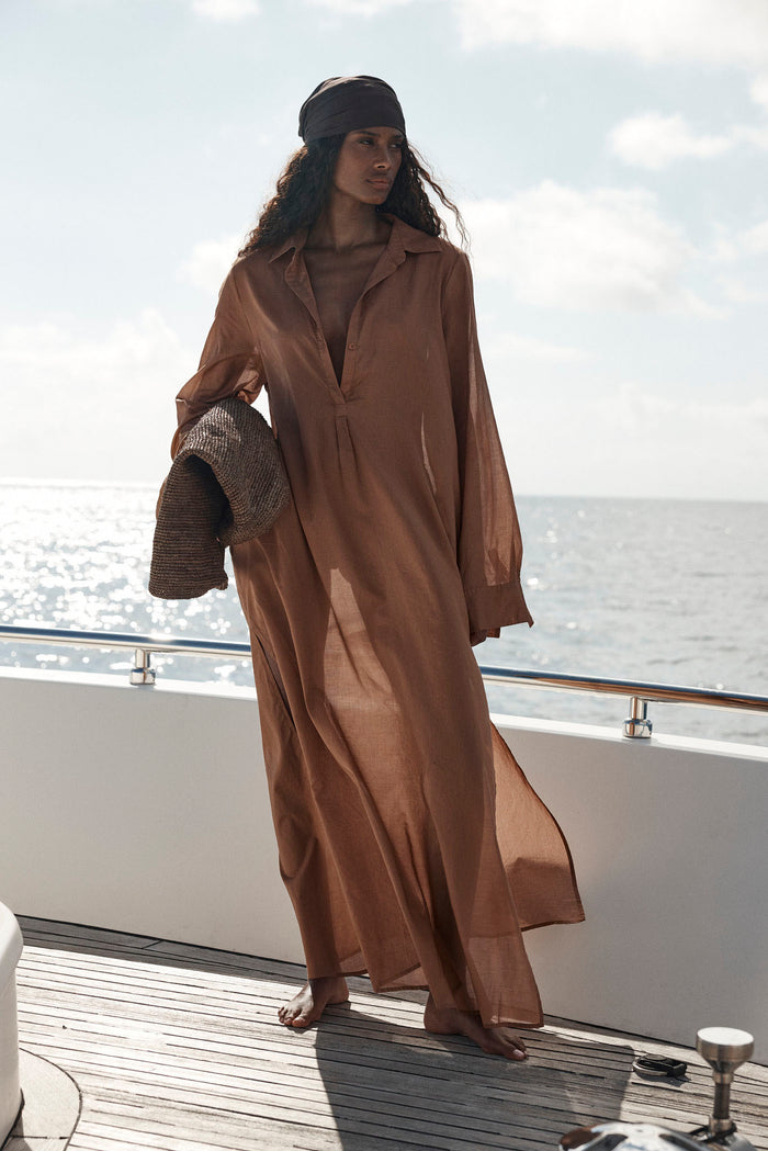 Woman wearing caramel Kokomo Kaftan in a loose fit standing on a boat deck at sea holding a woven sun hat