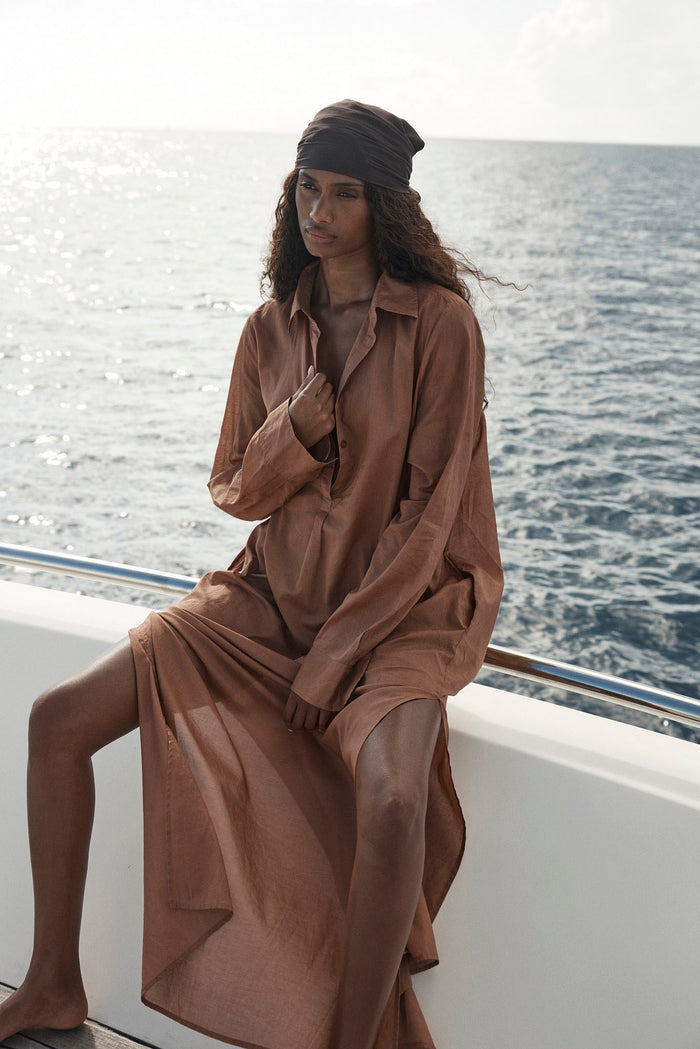Seated view of Kokomo Kaftan in caramel cotton voile showing button front, long sleeves and side slit on a boat by the ocean