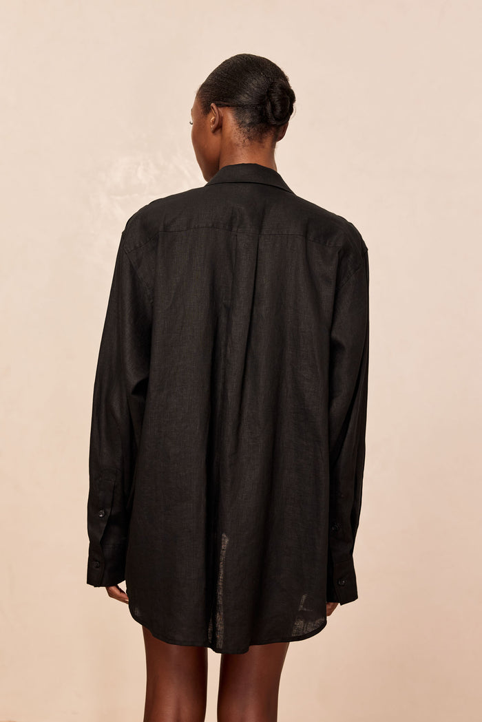 Back view of La Jolla Linen Shirt in black showing relaxed oversized fit, long sleeves, and curved hem on model against beige background