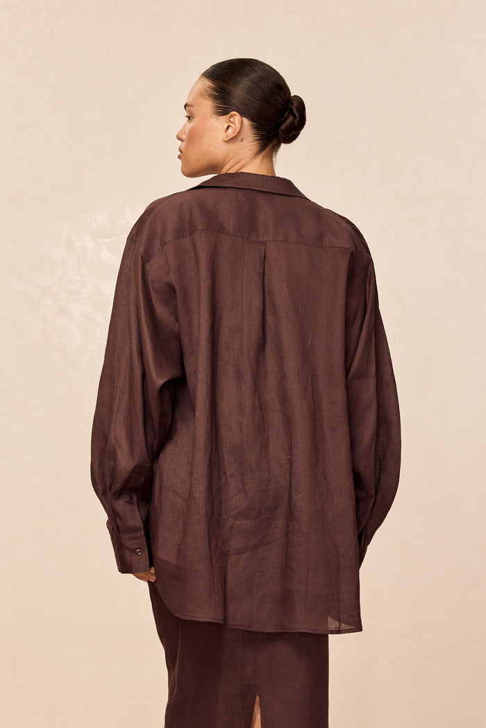 Back view of La Jolla linen shirt in espresso showing relaxed oversized fit, center back pleat and button cuffs on beige background