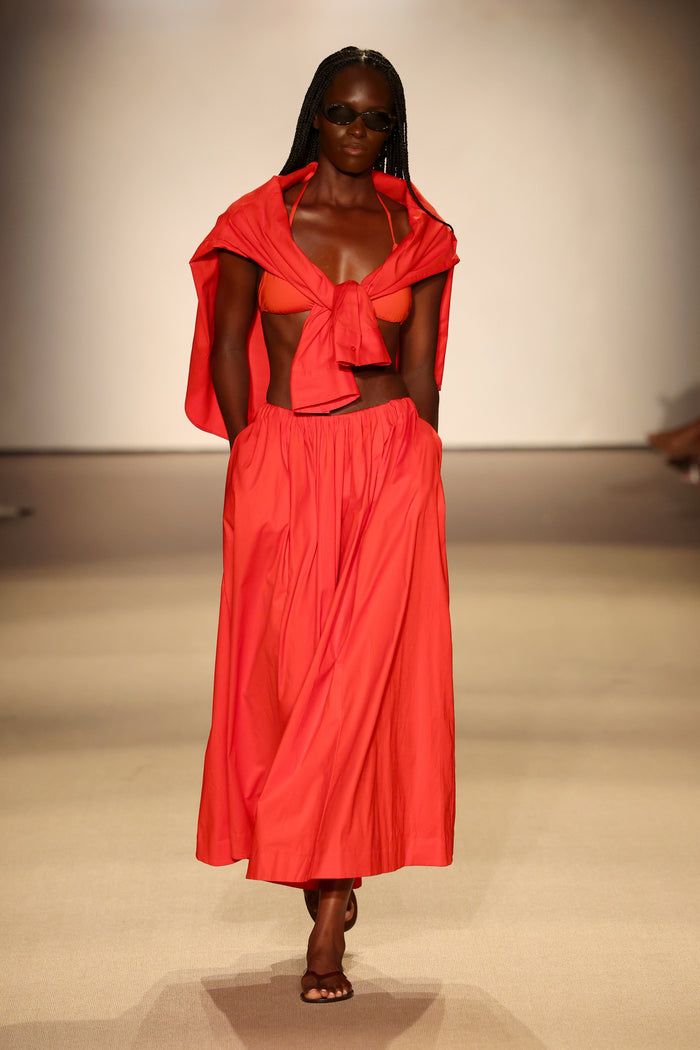 Runway model walking in Chili Pepper triangle bikini top and matching full skirt, shirt tied across shoulders and sunglasses against neutral background