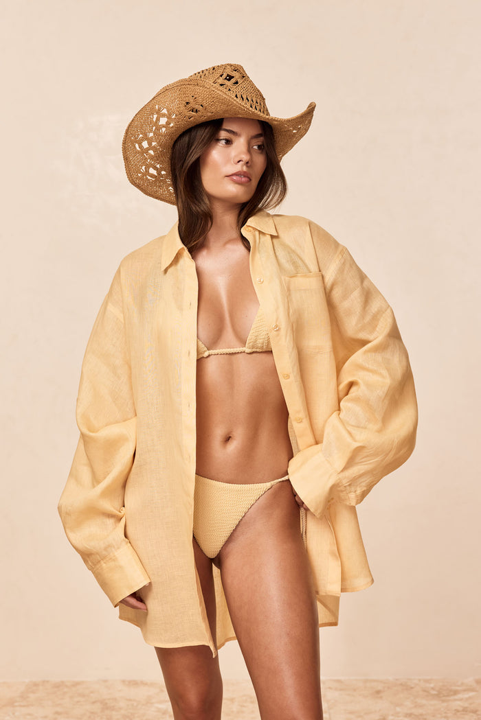 Three-quarter view of La Jolla Shirt in raffia linen, worn open over matching bikini to show crinkled texture and relaxed oversized fit on a neutral beige background