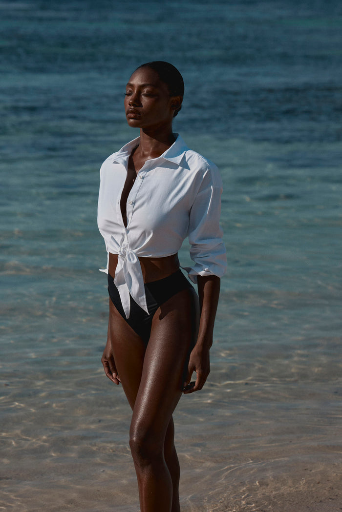 Front view of La Jolla Tie Shirt in white tied at front, semi-sheer long sleeves, modeled at ocean shoreline