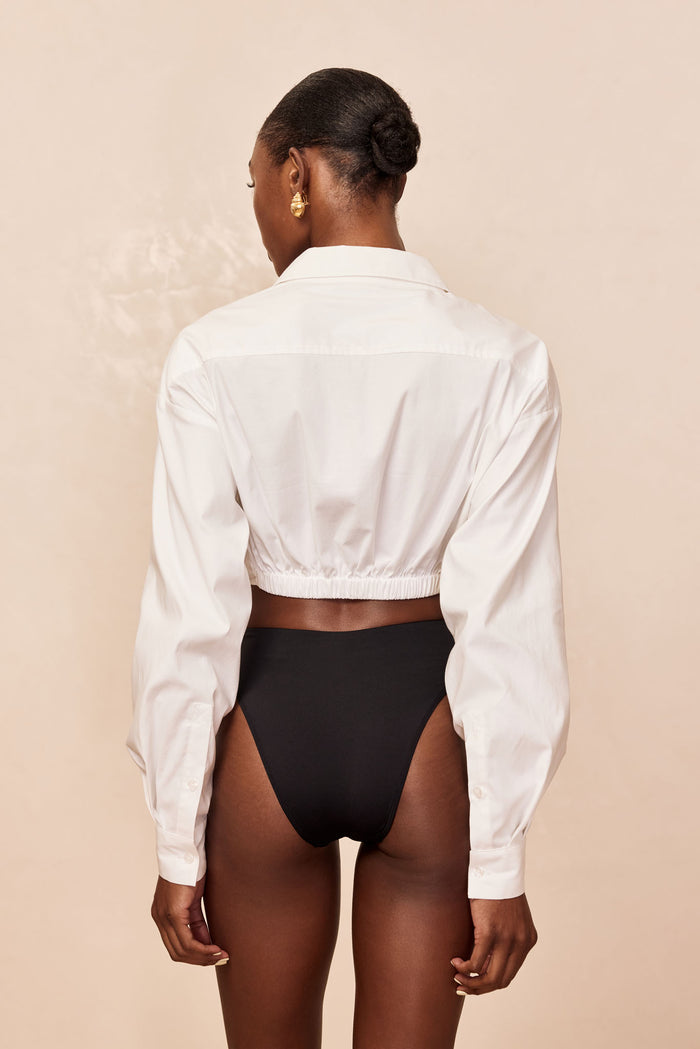 Back view of La Jolla Tie Shirt in white, cropped with elasticized hem, long sleeves and collar on beige background