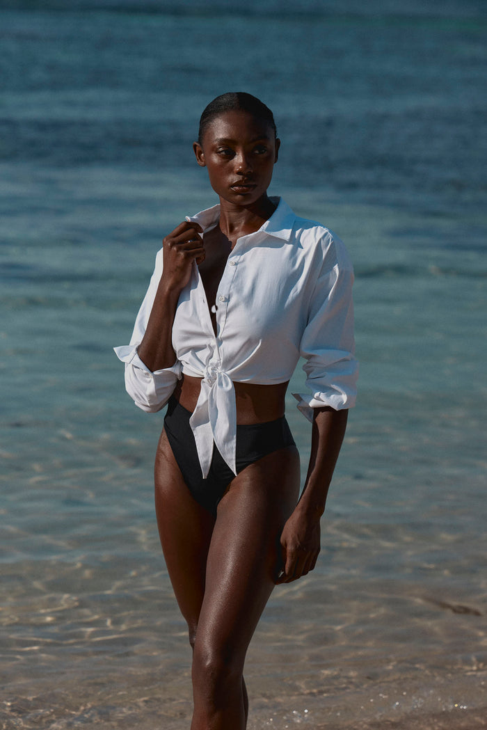 Front three-quarter view of La Jolla Tie Shirt in white, tied at the front to show cropped, semi-sheer long sleeves on a model at the beach
