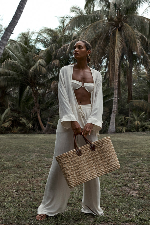 Three-quarter view of Lake Como Woven Bag Large showing hand-woven seagrass body and leather handles against palm trees
