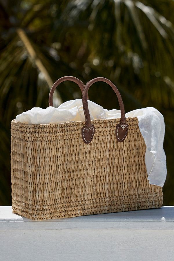 Three-quarter view of Lake Como Woven Bag Large showing seagrass weave, stitched brown leather handles, and white linen scarf against palm background