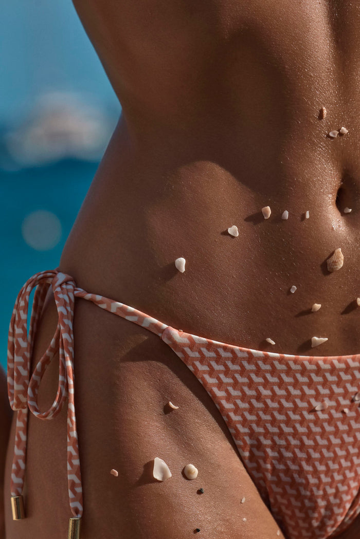 Close-up of Le Marais Bottom in Micro Geo showing adjustable tie sides and orange geometric print on model against blurred ocean