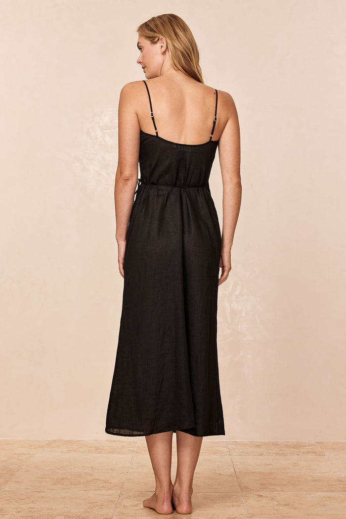 Back view of woman wearing a black midi dress with adjustable spaghetti straps and a tied waist, barefoot on a tile floor.