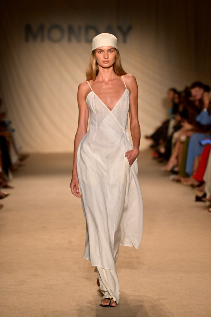 Front view of Levanzo Dress in white linen showcasing wrap V-neck, thin adjustable straps and tie waist with ‘MONDAY’ runway backdrop