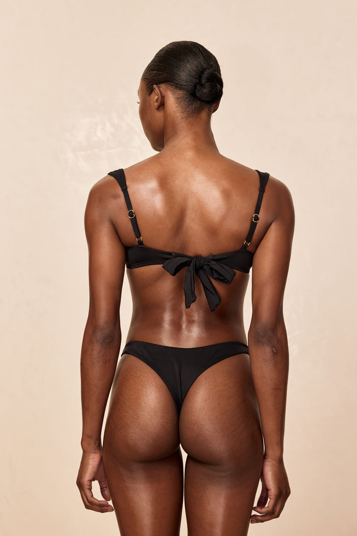 Rear view of Byron Thong in black on model showing high-cut minimal-coverage thong and tied-back bow against beige background