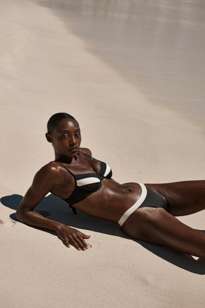 Reclining three-quarter view of Malta Top in Black/Ivory on model showing underwire cups and ivory contrast trim on sunlit sand