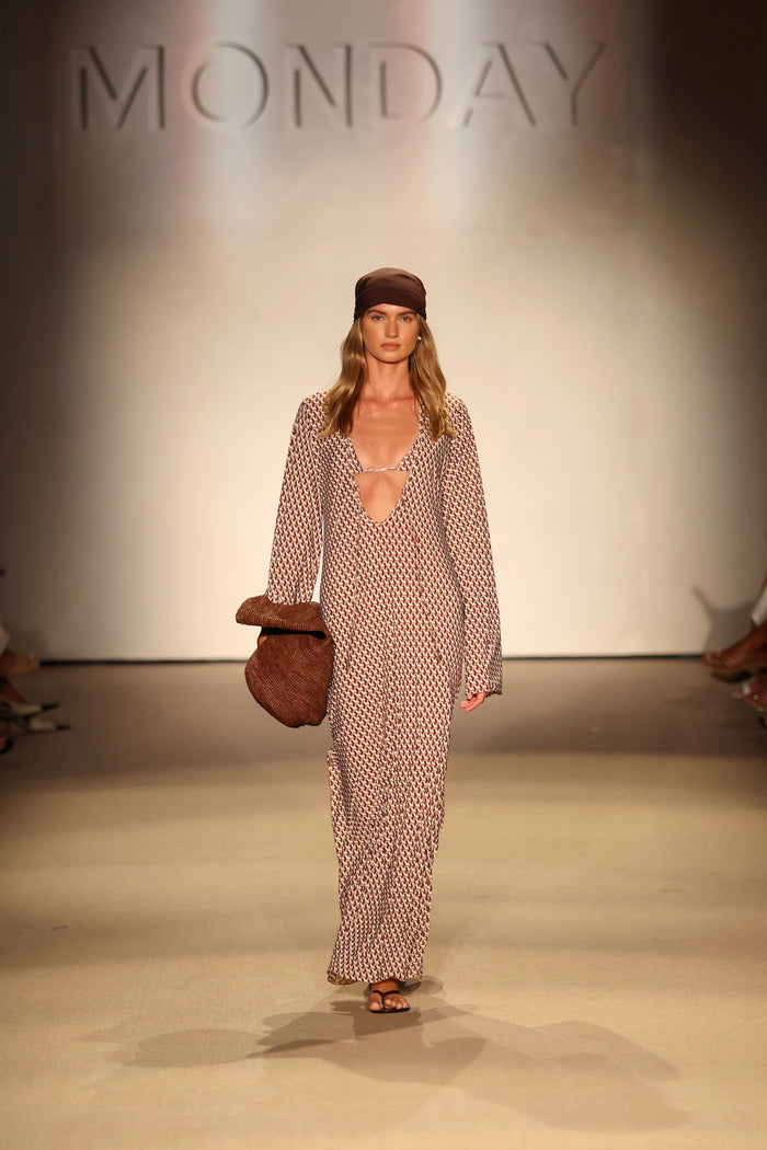Model walking runway wearing Marabella kaftan in husk geo print with deep V neckline, brown headscarf, woven tote, MONDAY backdrop