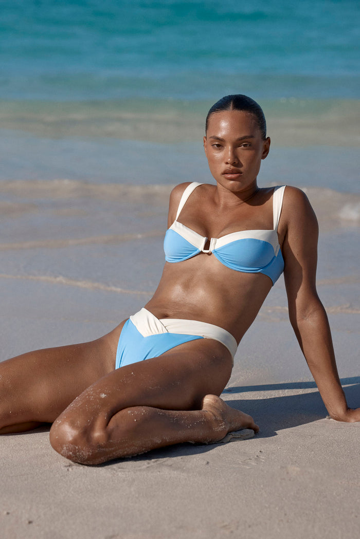 Seated three quarter view of Marseille Bottom in azure and ivory showing V-front high-rise waistband and moderate coverage on a model at the beach
