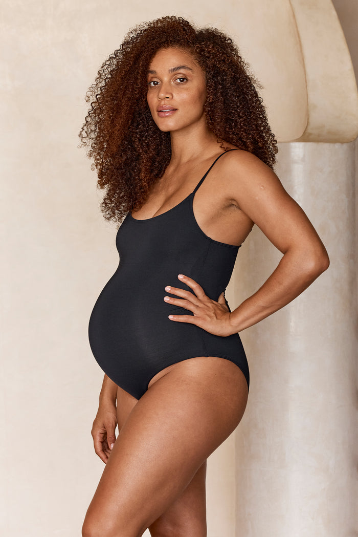 Side view of Maternity Bahamas One Piece in Black showing scooped neckline, thin straps and high-leg fit on pregnant model