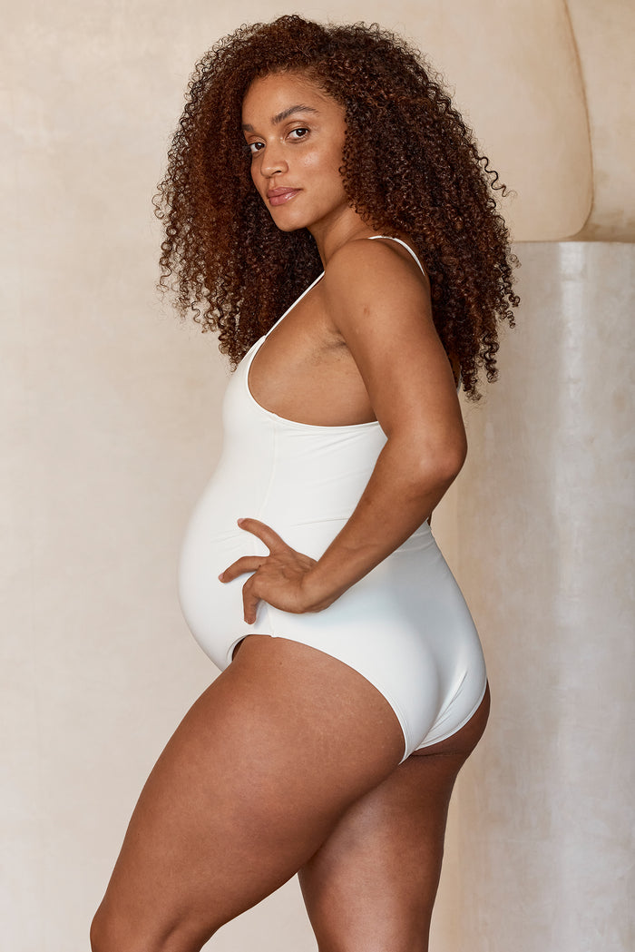 Side view of maternity Bahamas one-piece in ivory with high leg cut and scoop neckline on pregnant model, neutral background