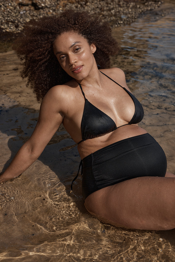 Three-quarter view of black Maternity Sorrento Bottom showing high-waisted fit and shirred front on pregnant model at beach