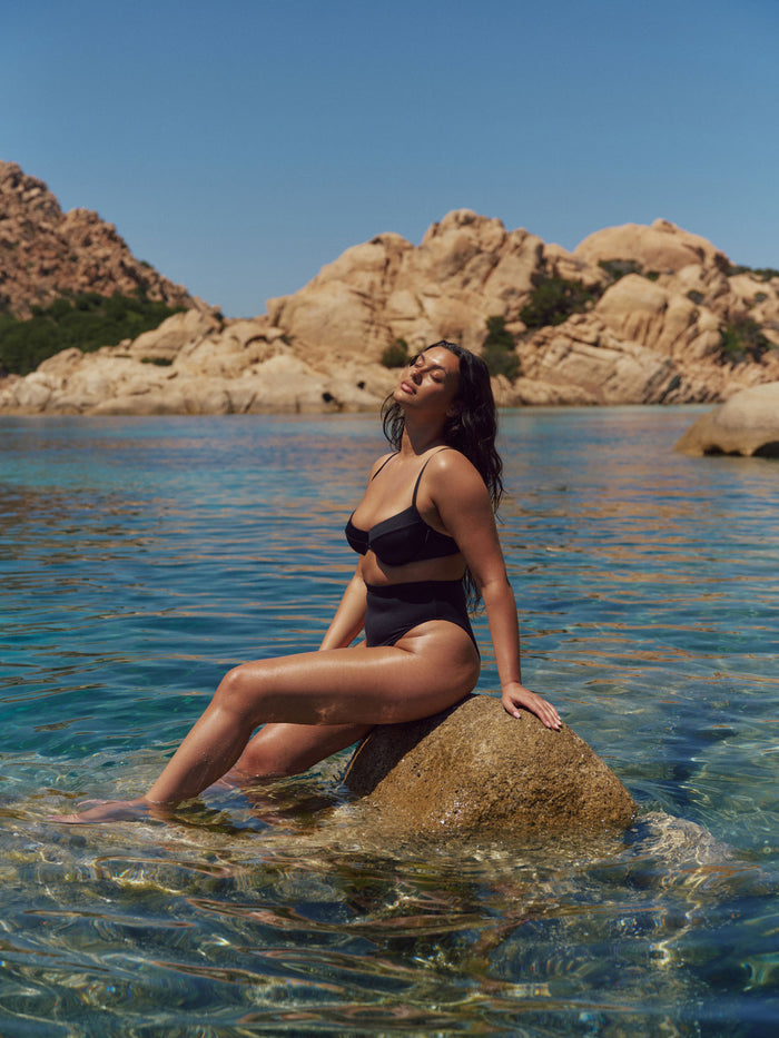 Woman wearing black Maui Top and high-rise bottoms, sitting on a rock in clear water with sunlit rocky coastline