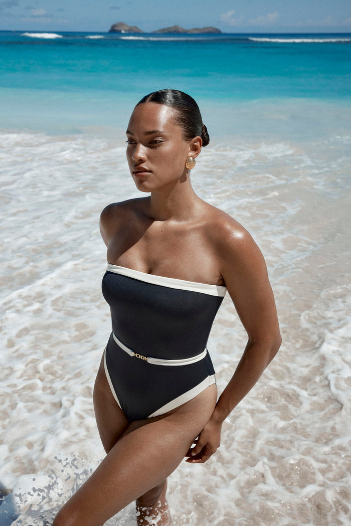 Three-quarter view of Miami One Piece black/ivory swimsuit showing bandeau neckline, gold MONDAY belt and beach background