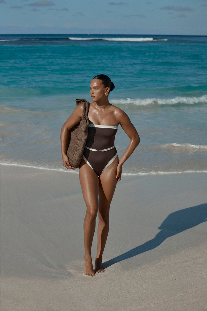 Model walking on a beach wearing espresso strapless high-cut one-piece with ivory trim and thin belt, carrying woven tote