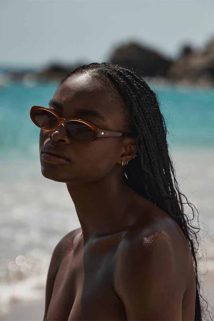 Three-quarter view of Milano sunglasses on model showcasing brown cat-eye acetate frame and gold rivet with sunlit beach background