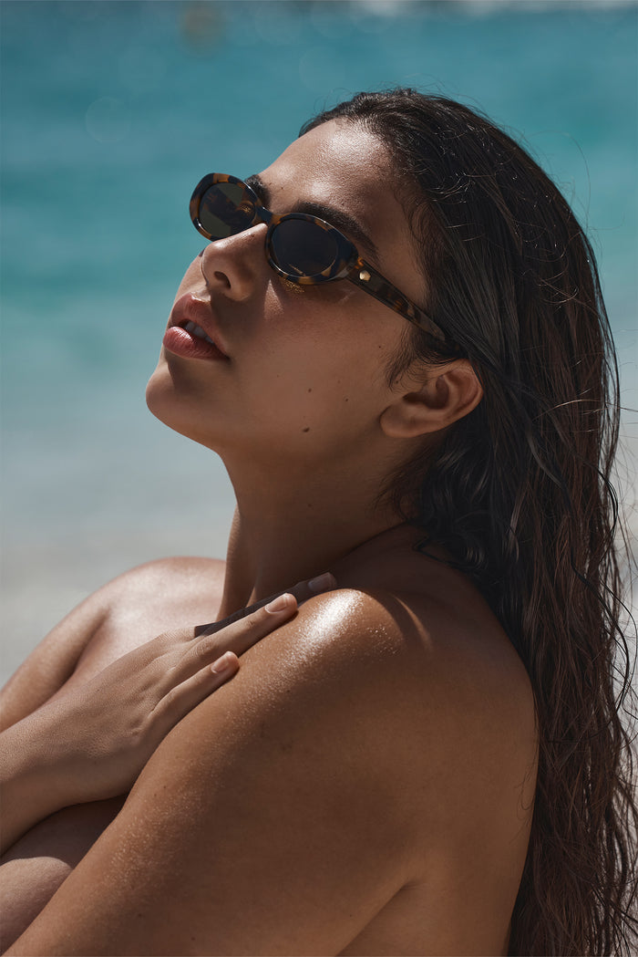Close-up of Milano sunglasses in tortoise showcasing oval frames and dark lenses on model with turquoise ocean background