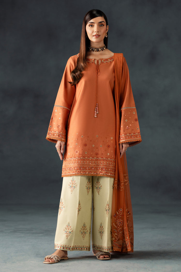 Woman wearing an orange embroidered outfit with beige pants against a dark background