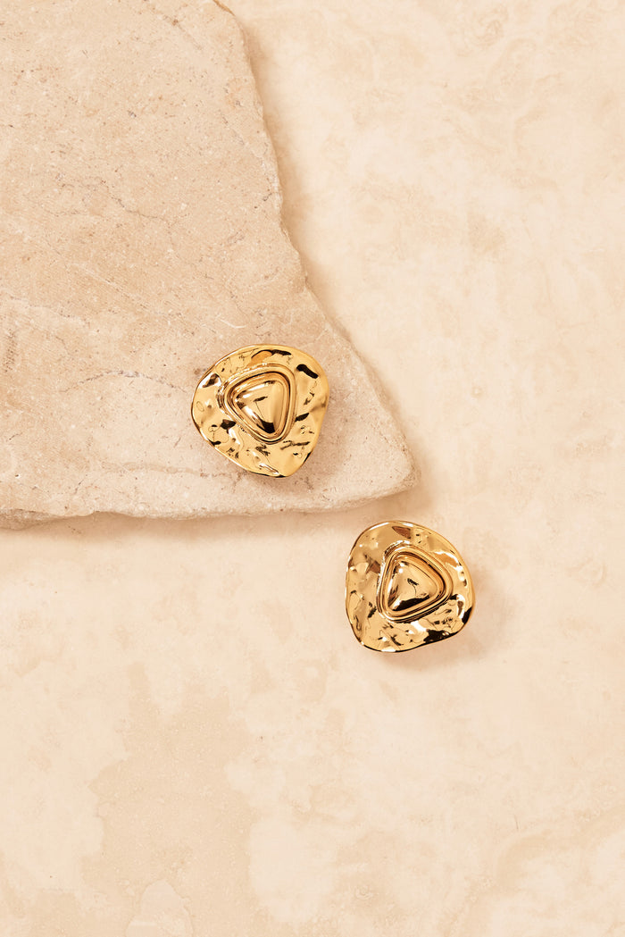 Top view of Monaco earrings in gold showcasing triangular 18K gold-plated sculptural shape with stud backing on beige stone background