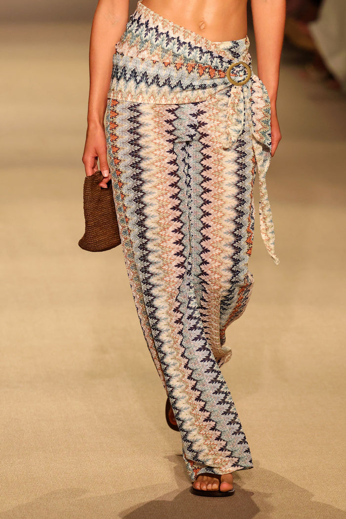 Model wearing printed sarong cinched at the hip with a gold Monday sarong buckle, holding a woven clutch on a runway