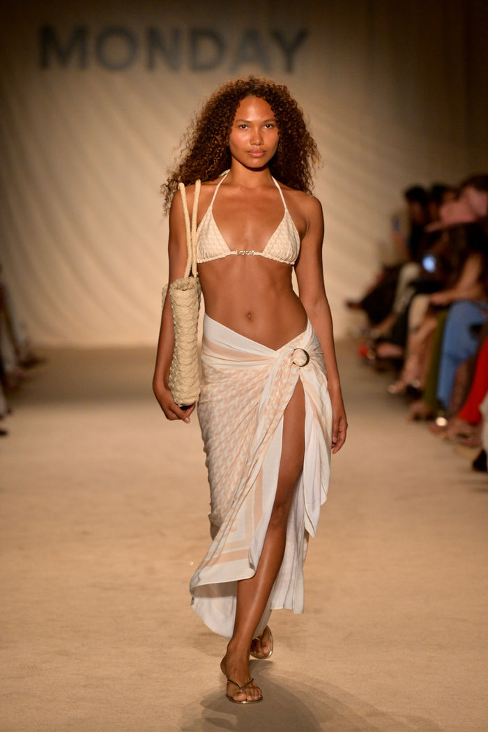 Model in cream bikini and sheer wrap skirt carrying a woven tote on runway; backdrop text reads MONDAY.
