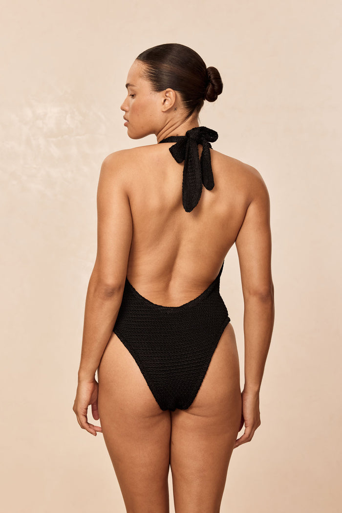 Back view of Montego Bodysuit in black treble knit showing low scooped back, halter tie and high-cut leg on beige background