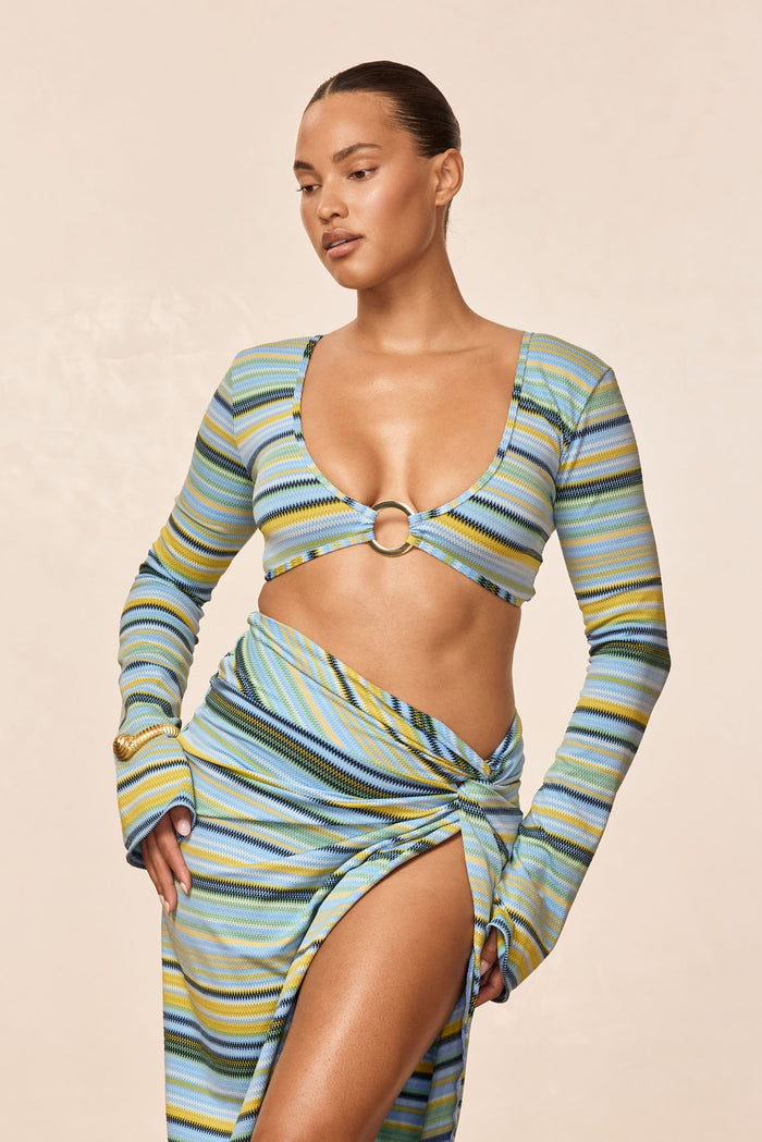 Front view of Montego crop top in Sardinia Stripe Azure with gold O-ring front, long sleeves on model against beige background