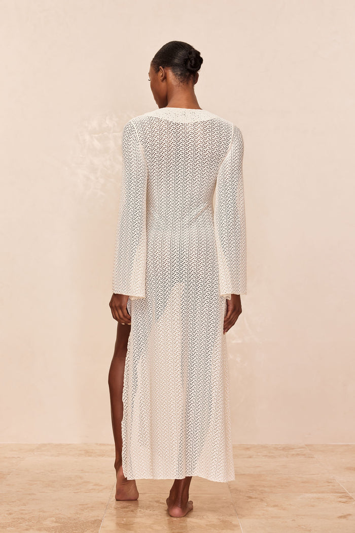 Rear view of Montego Dress in ivory crochet showing long sleeves and sheer maxi skirt with side slit on beige background