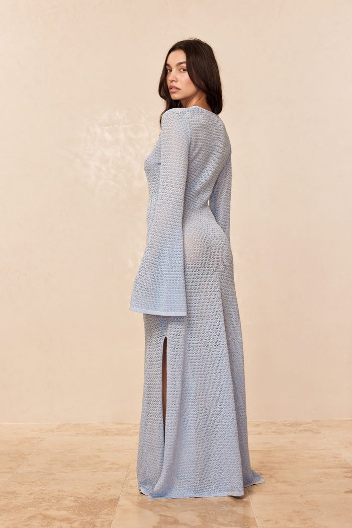 Rear three-quarter of Montego dress in sky lace crochet showing sheer knit, bell sleeves, and side slit on beige background