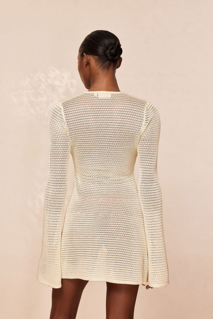 Back view of Montego Mini Dress in ivory shiny treble knit showing open-knit texture, fitted waist, and long flared sleeves on a neutral beige background
