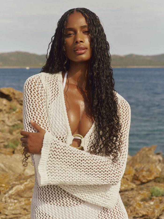 Front view of Montego Mini Dress in white petit crochet showing plunging neckline with gold ring and long flared sleeves by the sea