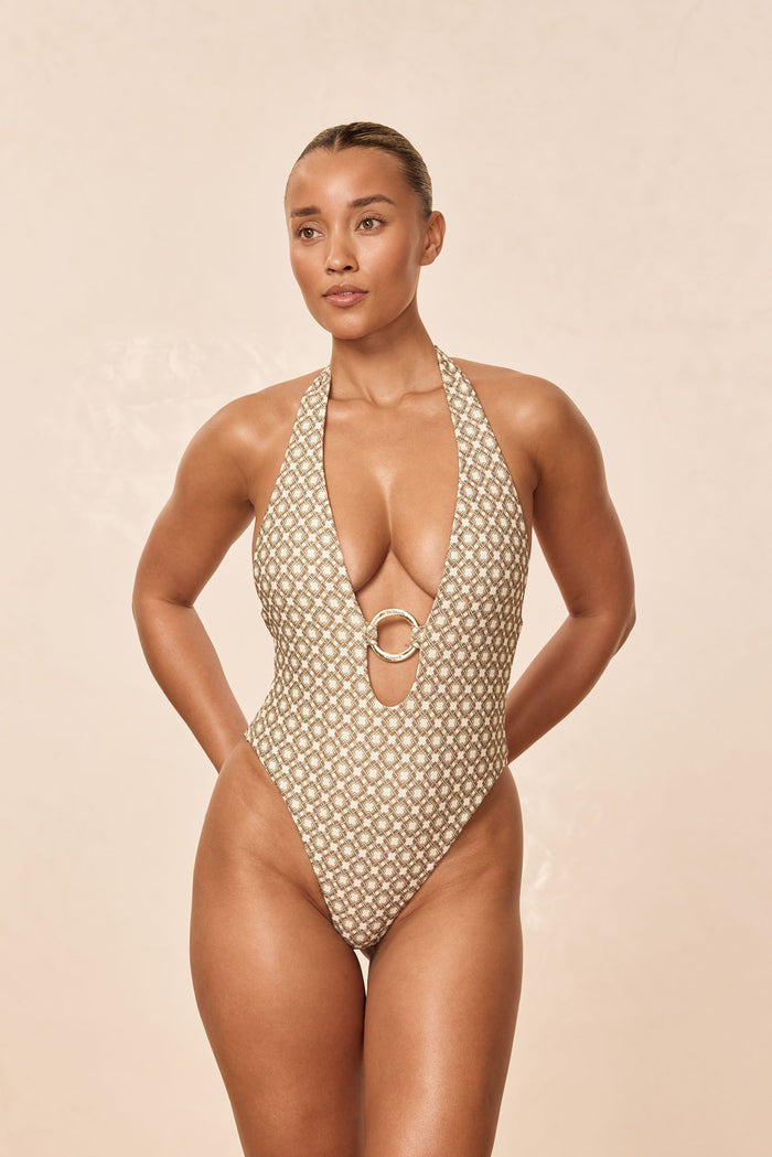 Front view of Montego Tile Geo one-piece swimsuit with deep halter plunge, gold O-ring, high-cut leg on beige background