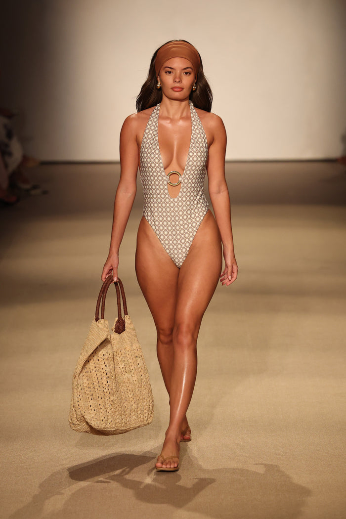 Model walking a beige runway wearing Montego One Piece in Tile Geo print with halter plunge, gold O-ring, high-leg cut and cheeky coverage