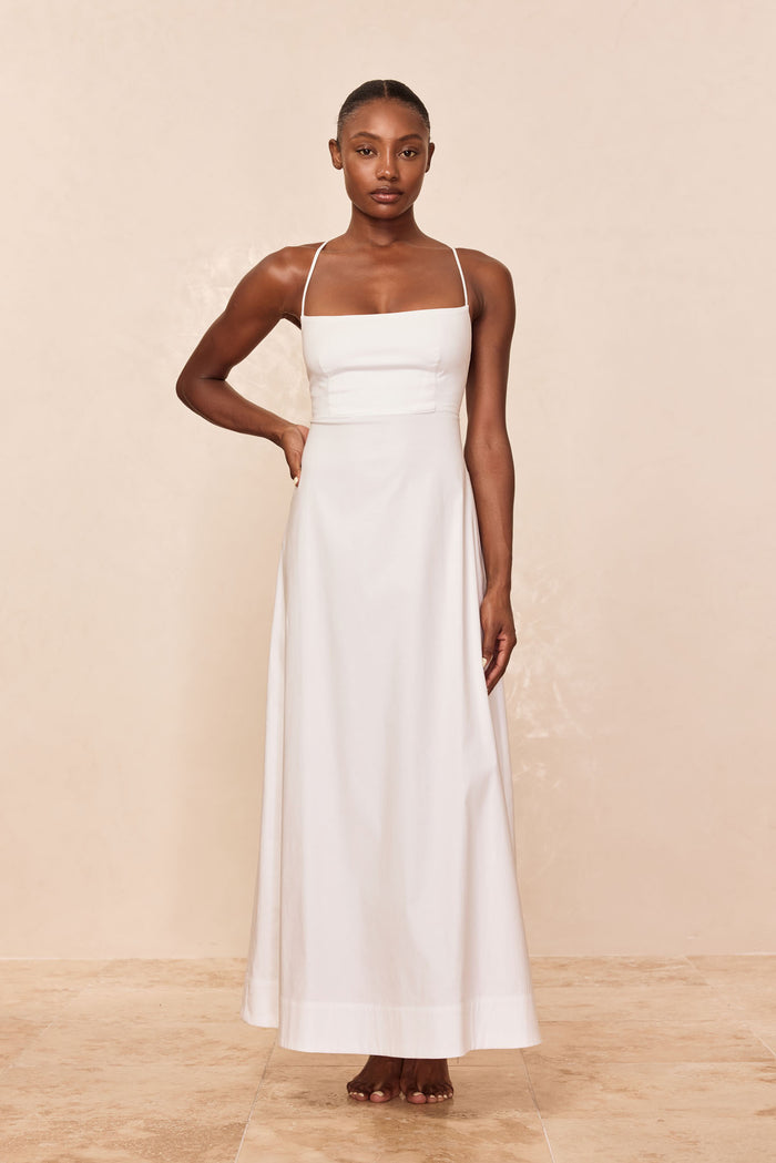 Model wearing a white spaghetti-strap maxi dress with square neckline, barefoot, one hand on hip, beige background