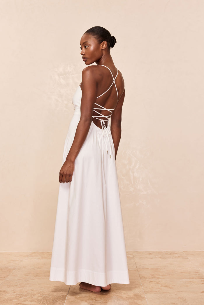 Model wearing long white backless dress with crisscross lace-up straps, shown from back, standing barefoot on tiled floor.