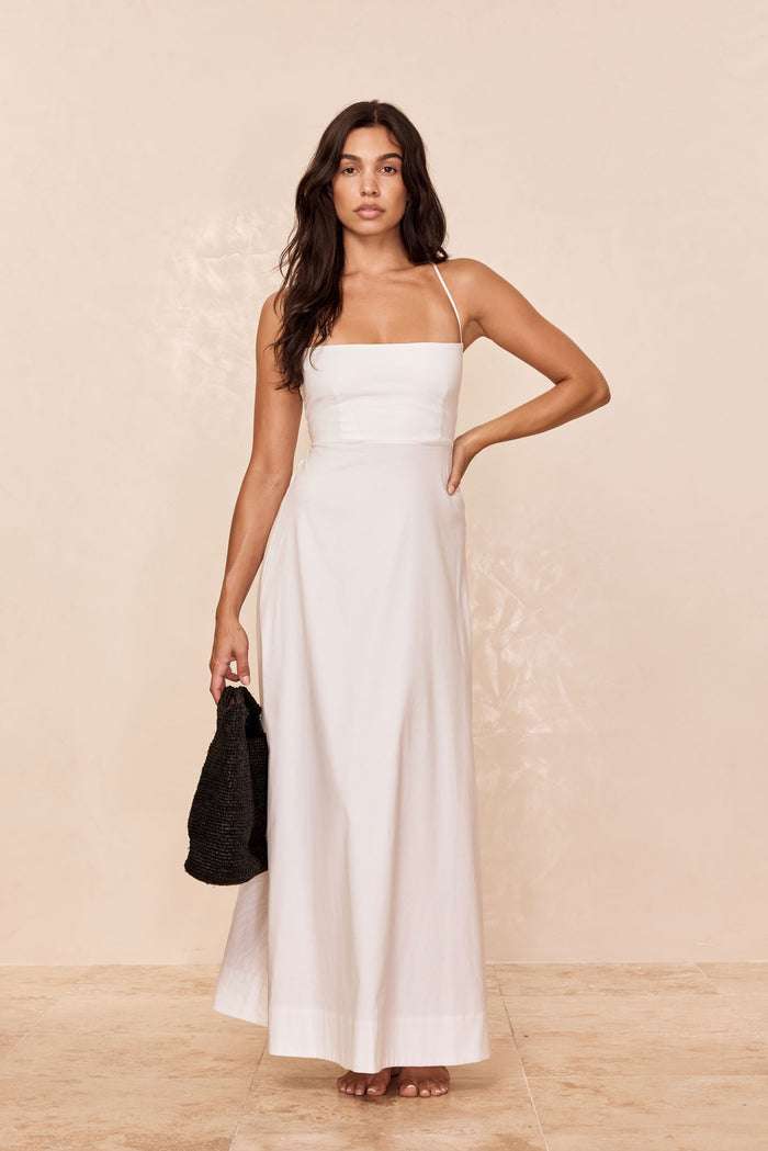 Woman modeling a white spaghetti-strap maxi dress, barefoot, hand on hip, holding a black woven bag.