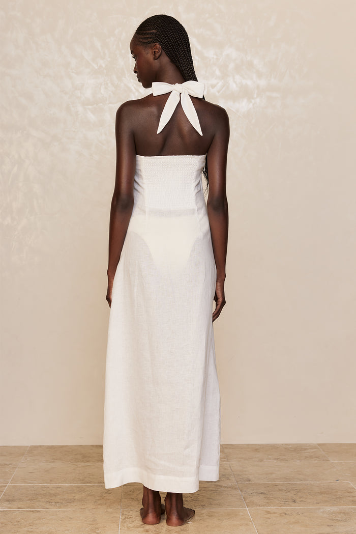 Model wearing a white strapless linen halter dress seen from the back; tied bow at the nape and floor-length skirt.